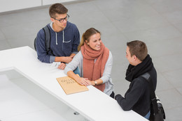 Sekretariat des Dekanats Drei Personen stehen neben einenander an einer Theke. Die Person in der Mitte lacht die Person rechts an und hat ihre Hände auf einen großen Umschlag liegen. Die Person links hat einen kleinen Zettel mit einer Wartenummer in der Hand.